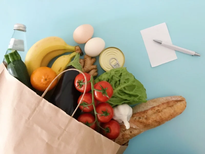 青い背景に置かれた紙袋から野菜や果物が見え、横にメモ帳とペンが置かれた食材の買い物シーン